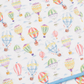A close-up of a white fabric item, likely a blanket or quilt, printed with a colorful watercolor-style hot air balloon pattern. A light blue stitched border is visible along the bottom edge.