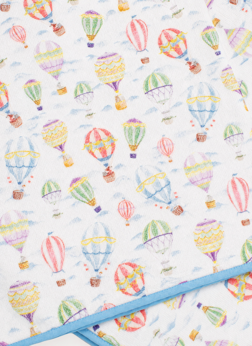 A close-up of a white fabric item, likely a blanket or quilt, printed with a colorful watercolor-style hot air balloon pattern. A light blue stitched border is visible along the bottom edge.