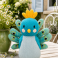 A knitted peacock toy bird sits on a wooden table outdoors, with a green pot of daisies and a garden background.