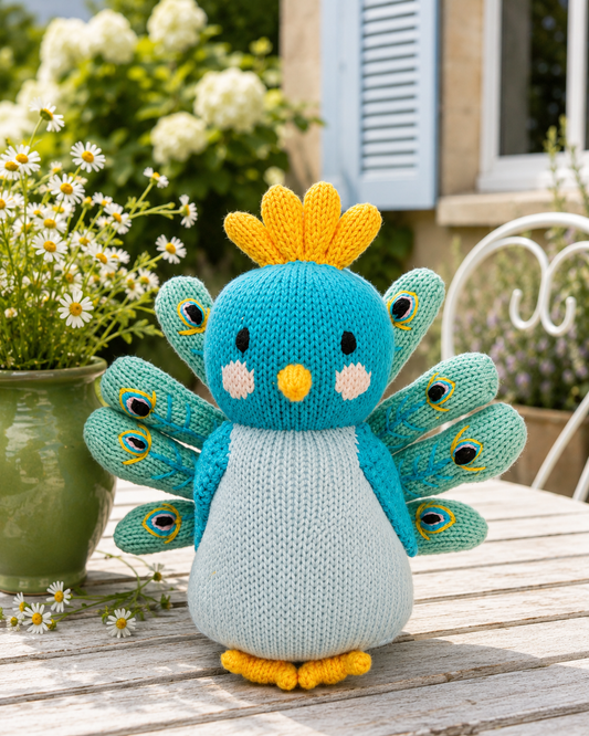 A knitted peacock toy bird sits on a wooden table outdoors, with a green pot of daisies and a garden background.