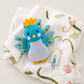 A small knitted peacock toy in bright blue, light blue, yellow, and orange, placed on top of a white muslin cloth printed with green vines, flowers, and a birdhouse.