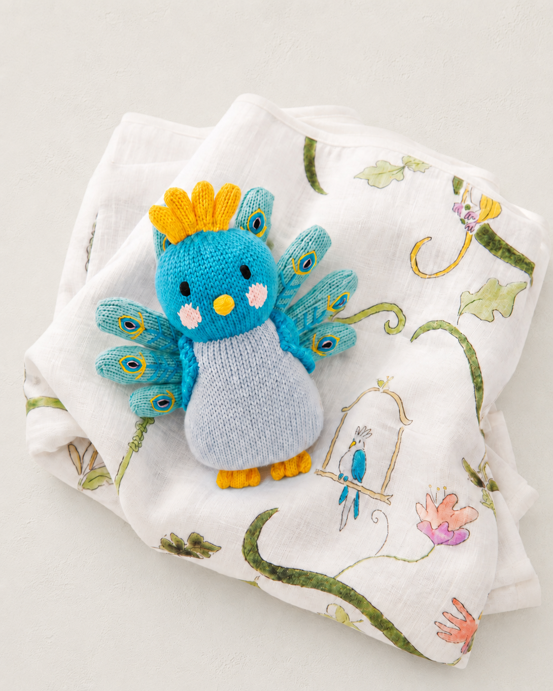 A small knitted peacock toy in bright blue, light blue, yellow, and orange, placed on top of a white muslin cloth printed with green vines, flowers, and a birdhouse.