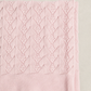 A folded light pink knit blanket with a textured cable-knit pattern, shown on a neutral beige background.