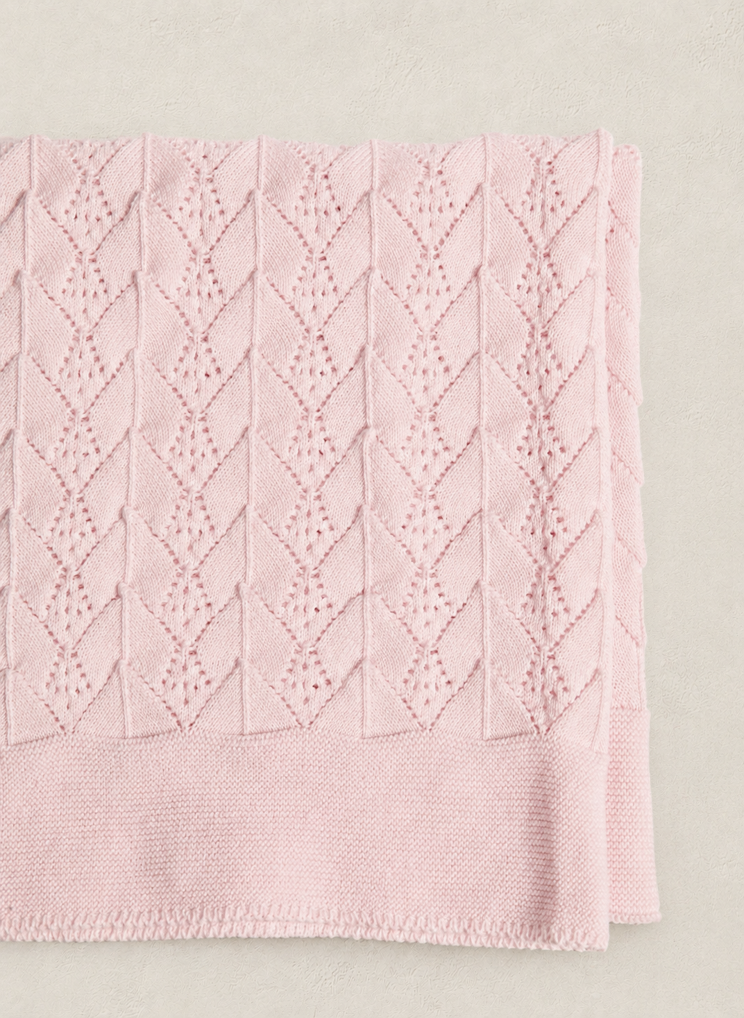 A folded light pink knit blanket with a textured cable-knit pattern, shown on a neutral beige background.