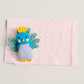 A small knitted peacock baby toy in blue, yellow, and gray, placed on top of a light pink textured baby blanket against a neutral light gray background.