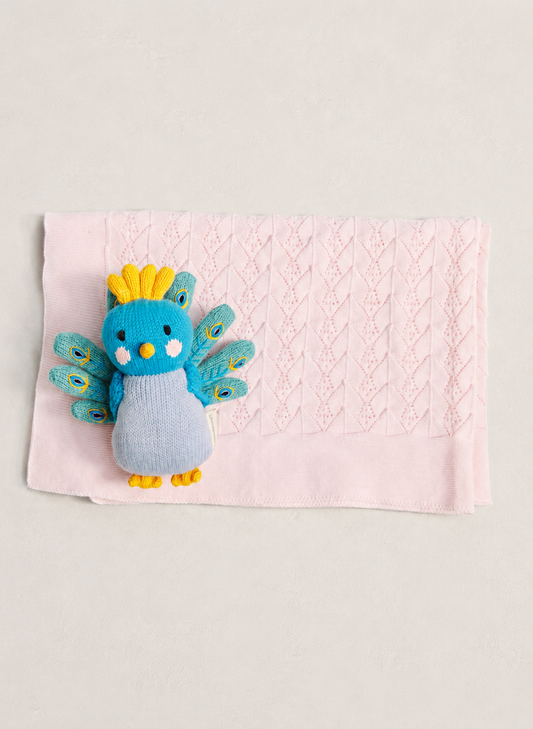 A small knitted peacock baby toy in blue, yellow, and gray, placed on top of a light pink textured baby blanket against a neutral light gray background.
