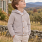 A young boy wearing a light beige textured knit hooded cardigan over a white collared shirt and beige pants, standing outdoors in a rural setting with a stone wall, house, and mountains in the background.