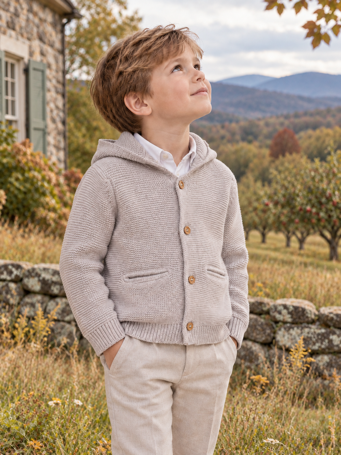 A young boy wearing a light beige textured knit hooded cardigan over a white collared shirt and beige pants, standing outdoors in a rural setting with a stone wall, house, and mountains in the background.