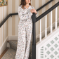 A woman wearing a matching long-sleeve pajama set with a delicate multicolor floral print on a white background, standing on a staircase landing next to a black banister.