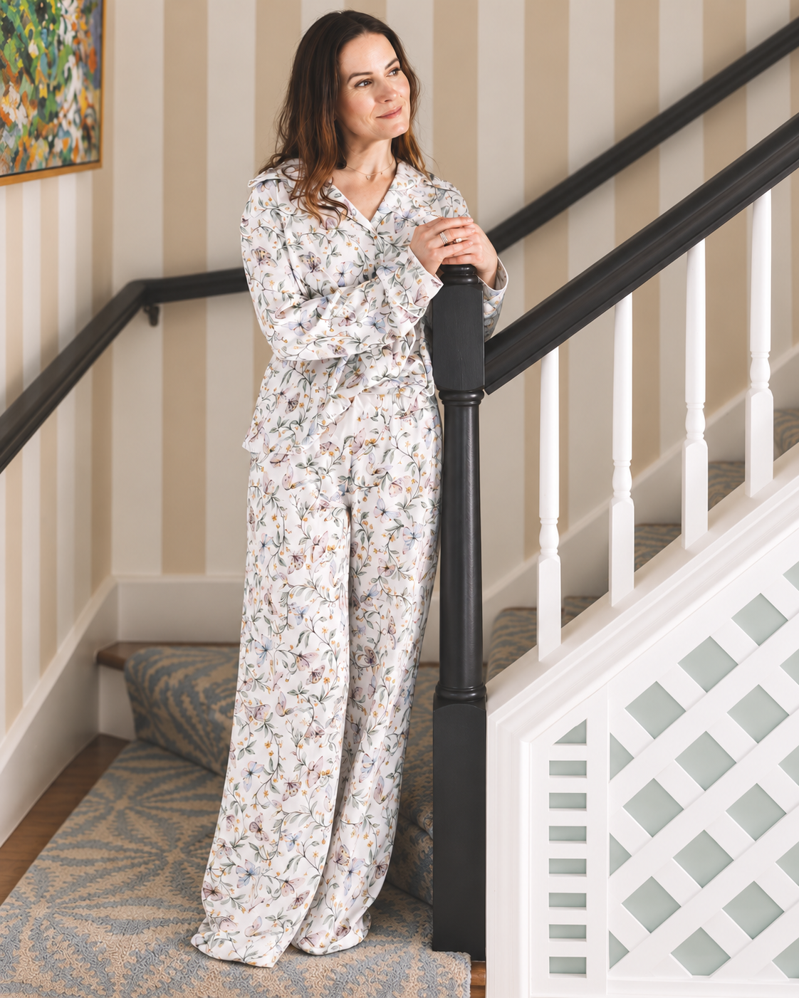 A woman wearing a matching long-sleeve pajama set with a delicate multicolor floral print on a white background, standing on a staircase landing next to a black banister.