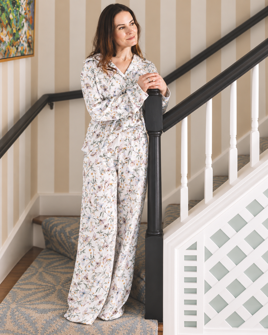 A woman wearing a matching long-sleeve pajama set with a delicate multicolor floral print on a white background, standing on a staircase landing next to a black banister.