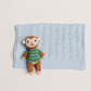 A light blue knitted baby blanket with a textured diamond pattern, styled flat-lay with a small knitted monkey toy wearing a green and blue striped sweater.