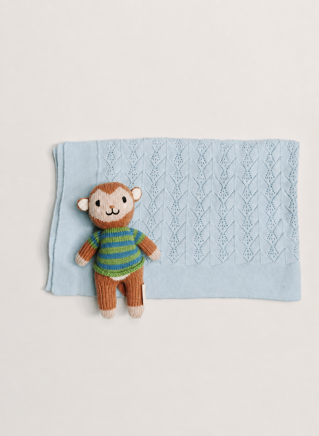 A light blue knitted baby blanket with a textured diamond pattern, styled flat-lay with a small knitted monkey toy wearing a green and blue striped sweater.