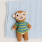 A knitted monkey toy wearing a green and blue striped sweater, posed on top of a light blue textured knit blanket against a warm off-white background.