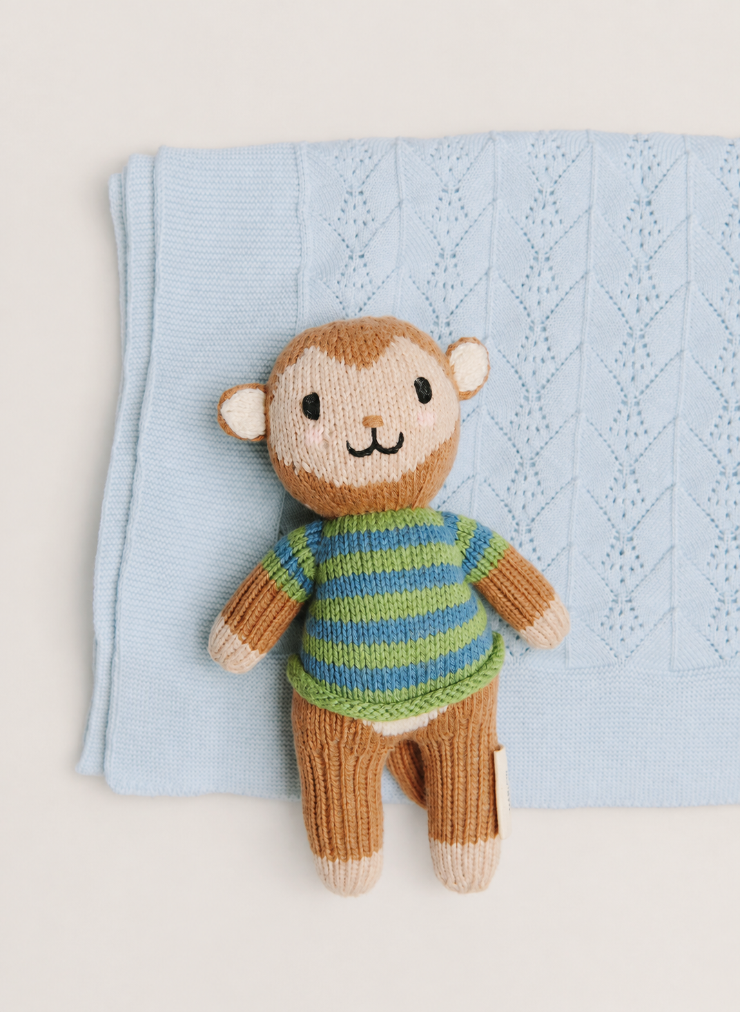 A knitted monkey toy wearing a green and blue striped sweater, posed on top of a light blue textured knit blanket against a warm off-white background.