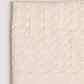 A folded cream-colored textured fabric, likely a throw blanket or knit blanket, featuring a raised diamond cable-knit pattern and a solid ribbed border, shown against a neutral light gray background.