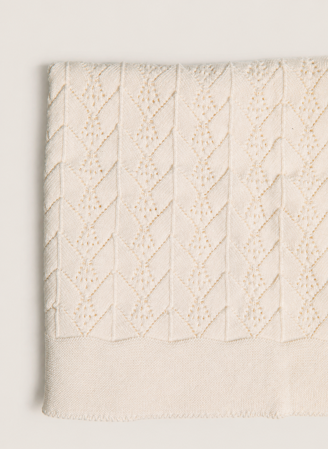 A folded cream-colored textured fabric, likely a throw blanket or knit blanket, featuring a raised diamond cable-knit pattern and a solid ribbed border, shown against a neutral light gray background.