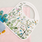 A white baby bib with colorful embroidered birds, flowers, and foliage, styled on a pink textured blanket with a blue knitted bootie prop against a light gray background.