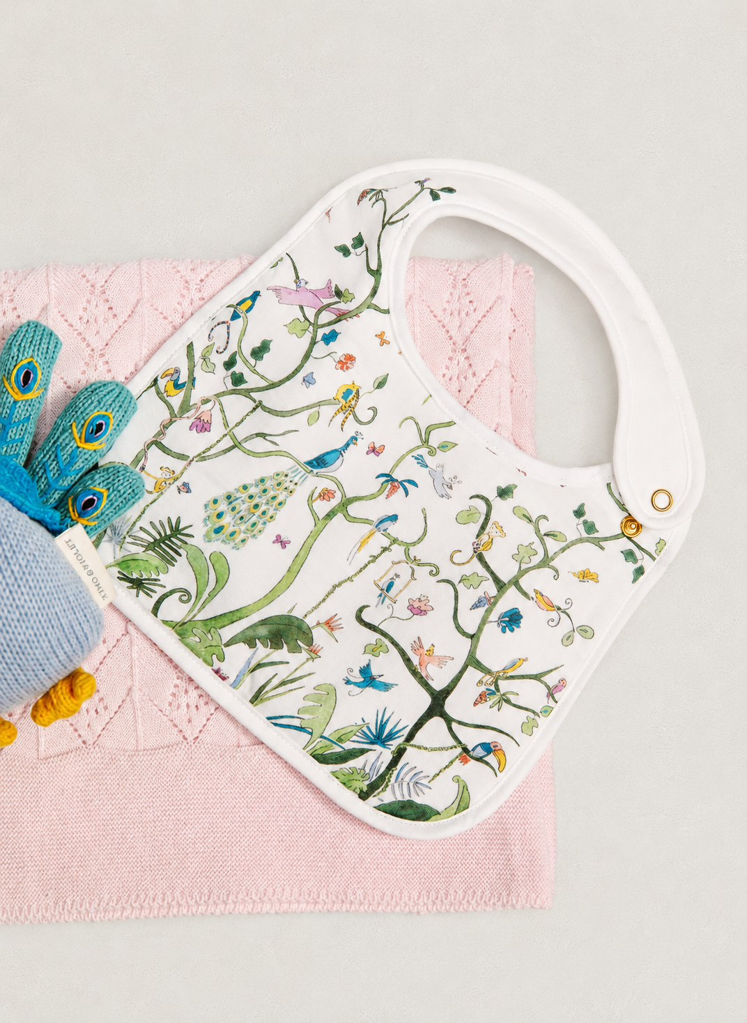 A white baby bib with colorful embroidered birds, flowers, and foliage, styled on a pink textured blanket with a blue knitted bootie prop against a light gray background.