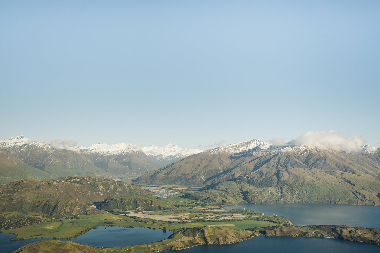 Scenic aerial view of green valleys, blue lakes, and snow-capped mountains under a clear blue sky.