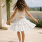 A young girl with long brown hair is seen from behind, twirling outdoors on a stone patio. She is wearing a white sleeveless top and a white tiered ruffle skirt decorated with a delicate pastel floral and butterfly print.