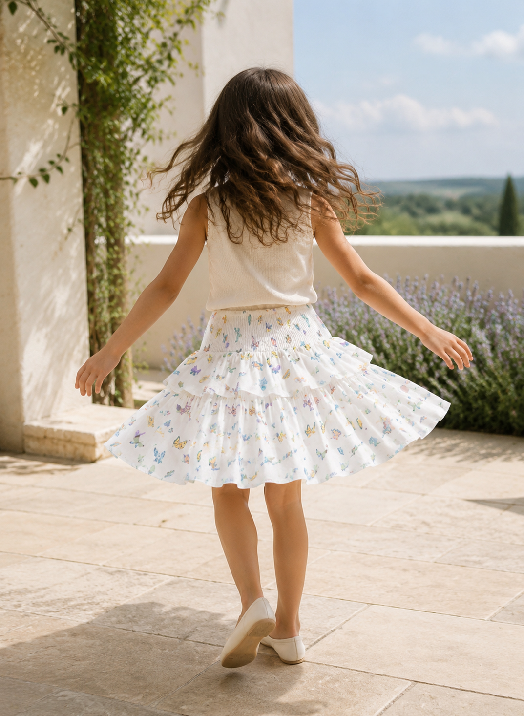 A young girl with long brown hair is seen from behind, twirling outdoors on a stone patio. She is wearing a white sleeveless top and a white tiered ruffle skirt decorated with a delicate pastel floral and butterfly print.