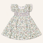A toddler girl's ruffled dress with a delicate multicolor floral print on a cream background, featuring a smocked waist and flutter sleeves, shown flat on a light beige background.