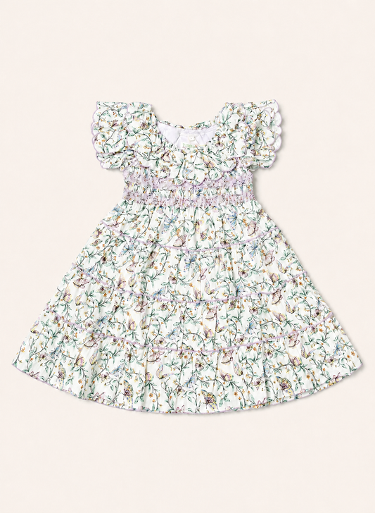 A toddler girl's dress with a white base and a delicate multicolor floral print, featuring ruffled cap sleeves and a smocked waist, shown flat on a light beige background.
