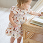 A toddler girl with blonde hair stands barefoot next to a rattan table, wearing a cream tiered dress with a delicate multicolor floral and bird pattern.