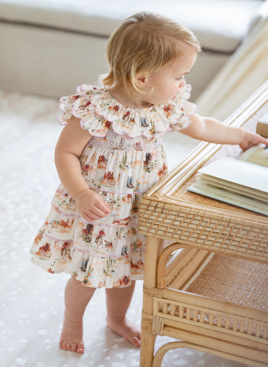 A toddler girl with blonde hair stands barefoot next to a rattan table, wearing a cream tiered dress with a delicate multicolor floral and bird pattern.