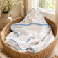 A baby hooded towel with a colorful hot air balloon print and blue trim, displayed inside a woven wicker basket on a wooden floor next to a vase of baby's breath.